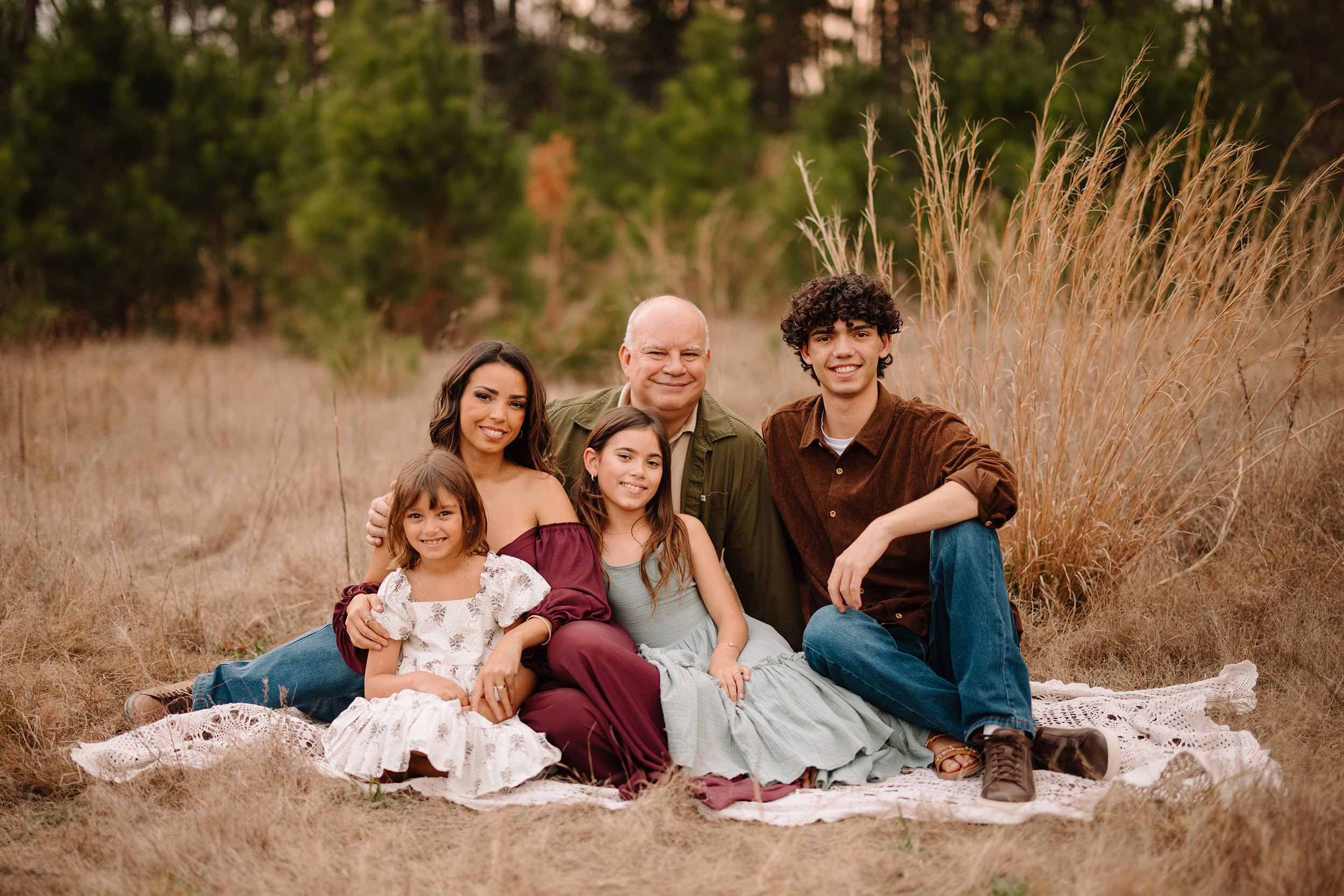 Family sitting on a blanket in a field of tall grass with trees in the background.