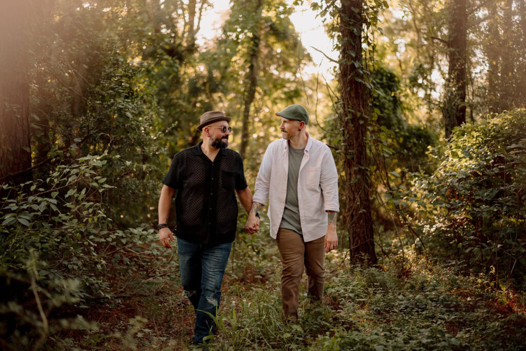Two men walking hand-in-hand through a sunlit forest trail.