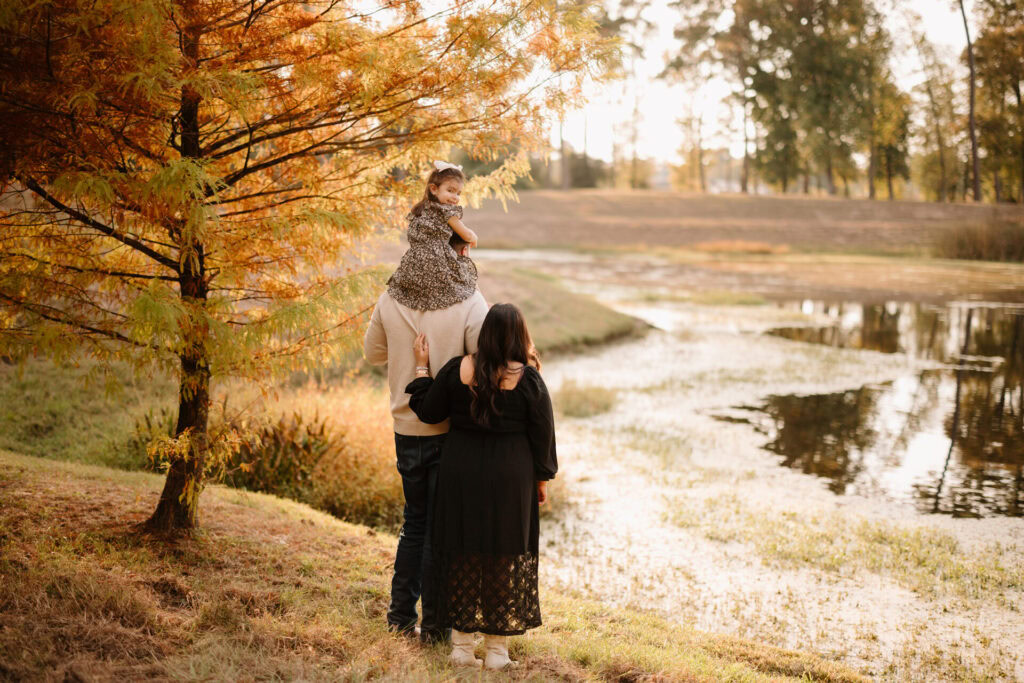 Autumn family photo with a child on dad's shoulders near a pond in The Woodlands, TX.