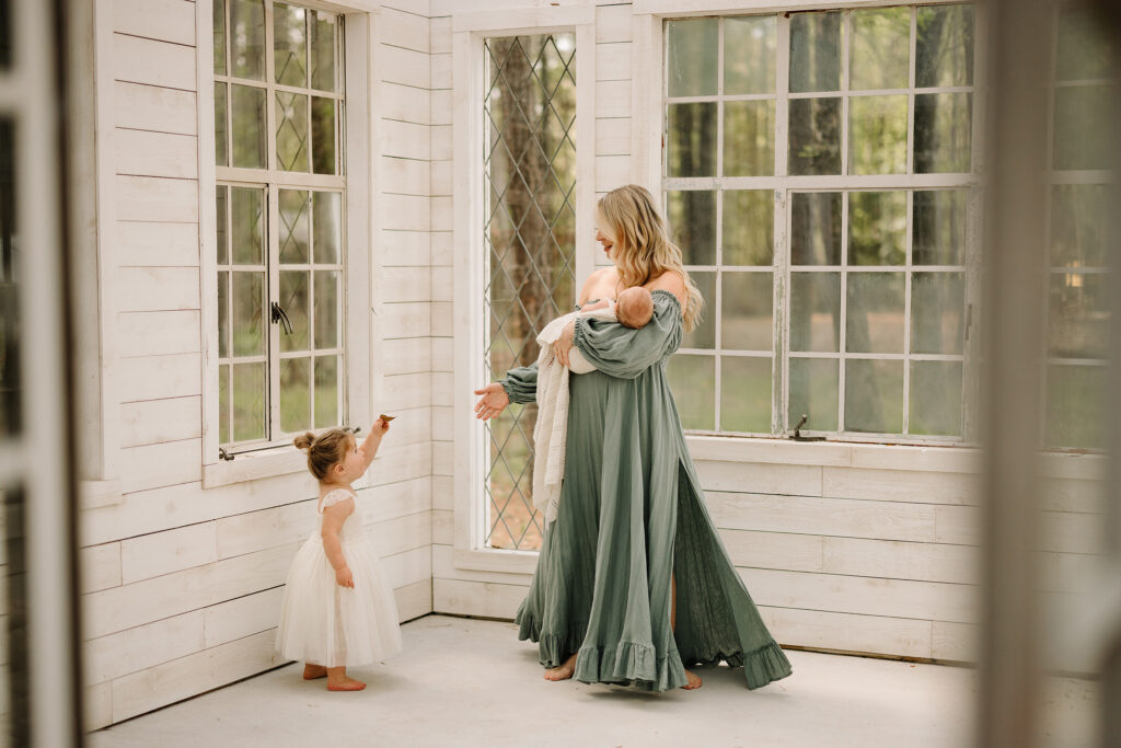 Mother with two children in a sunlit porch, holding a baby and playing with a young girl.