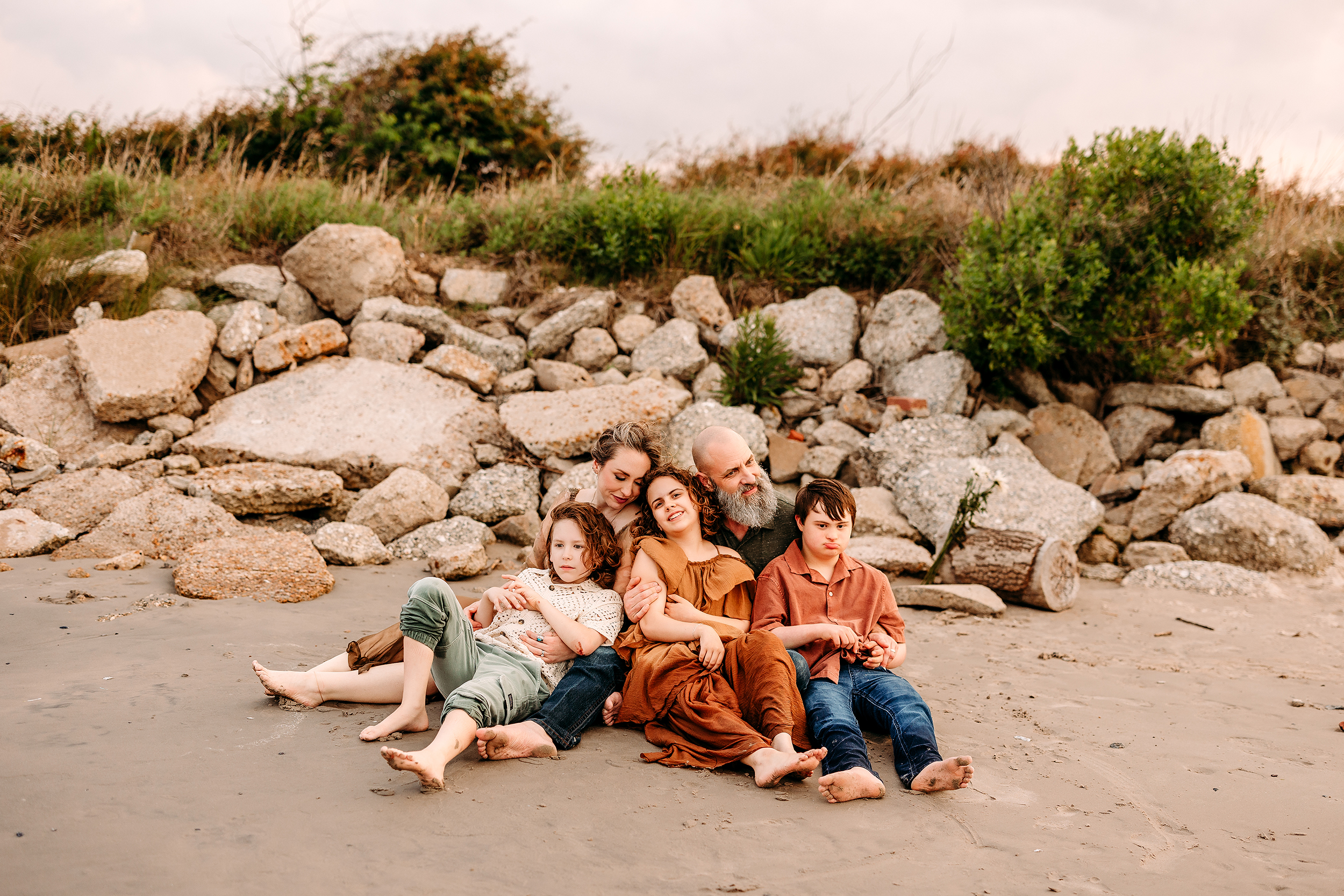 Family sitting on sandy beach with rocks and lush greenery in the background, enjoying a relaxed mom.