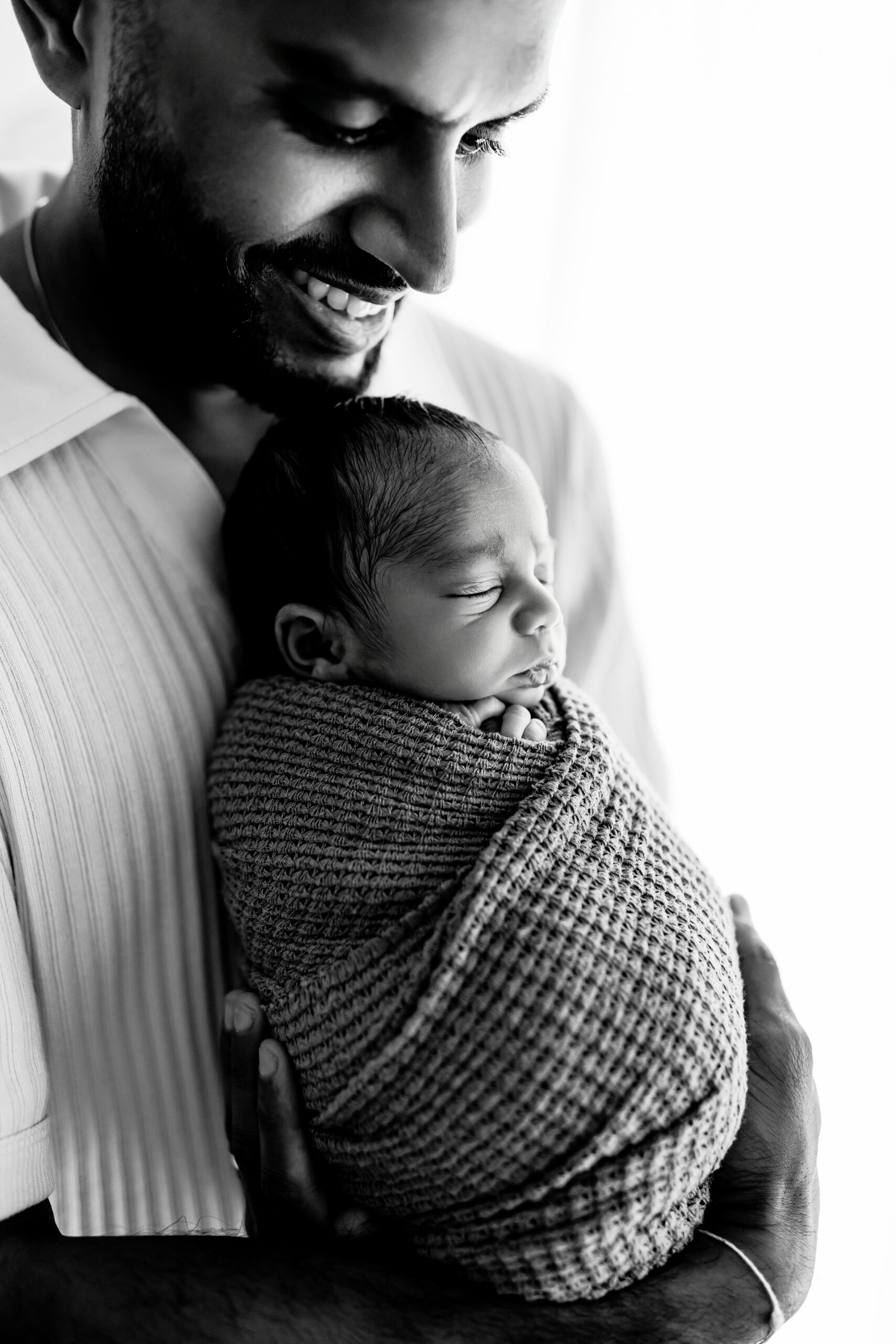Father holding sleeping newborn wrapped in blanket, smiling gently in black and white.