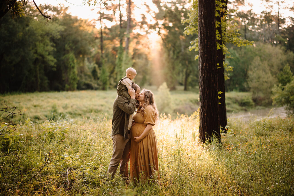 Family portrait of a pregnant woman, her partner, and their young child in a sunlit forest clearing.