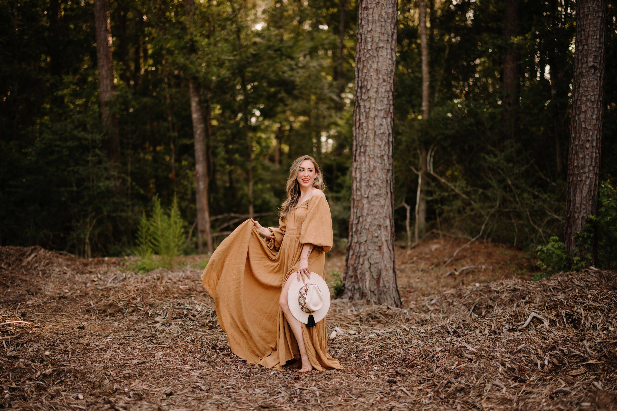 Woman in a flowing dress holding a hat in a wooded outdoor setting.